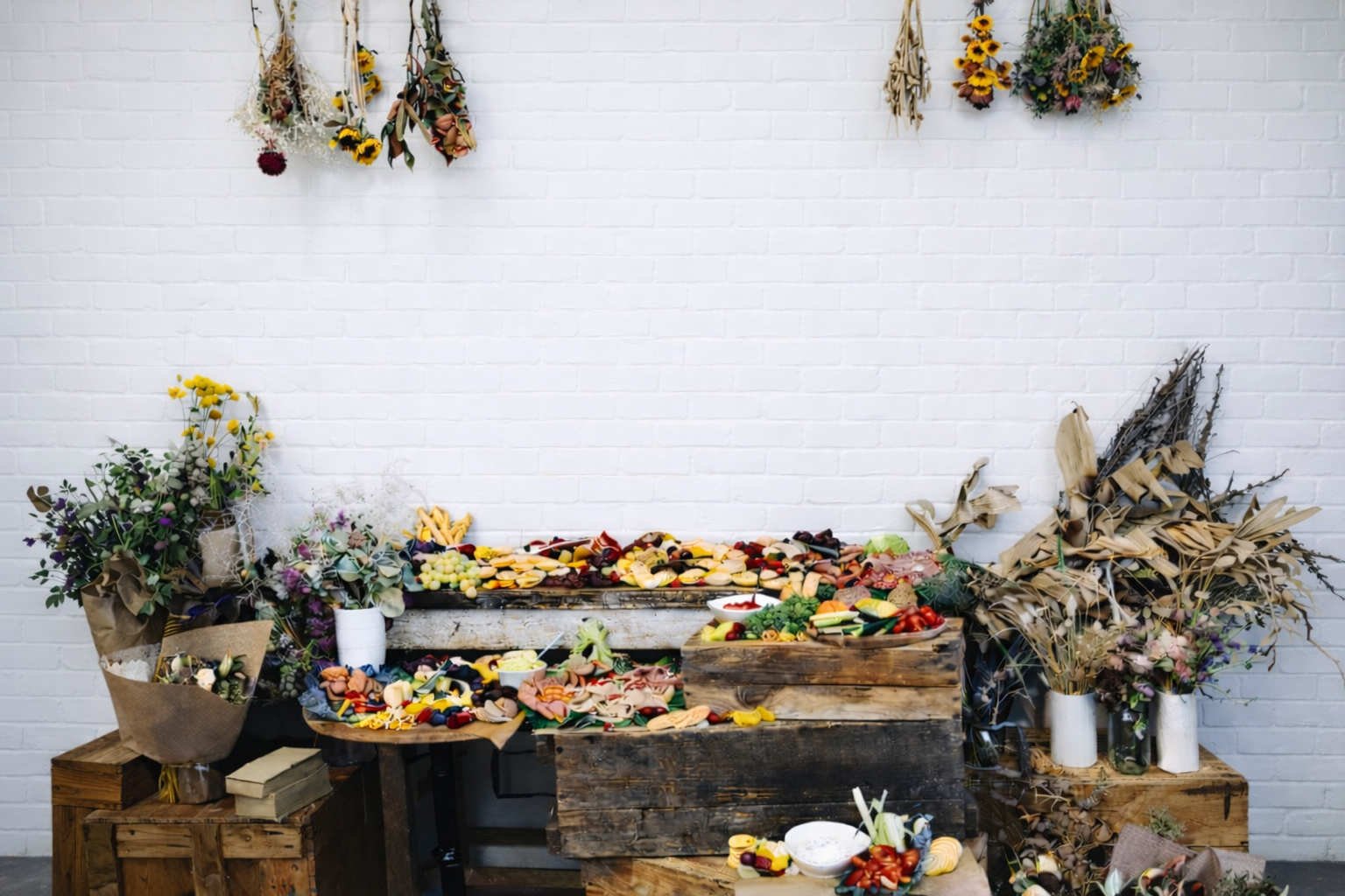Decorative grazing table setup with flowers, fruits, and vegetables on wooden crates against a white wall.